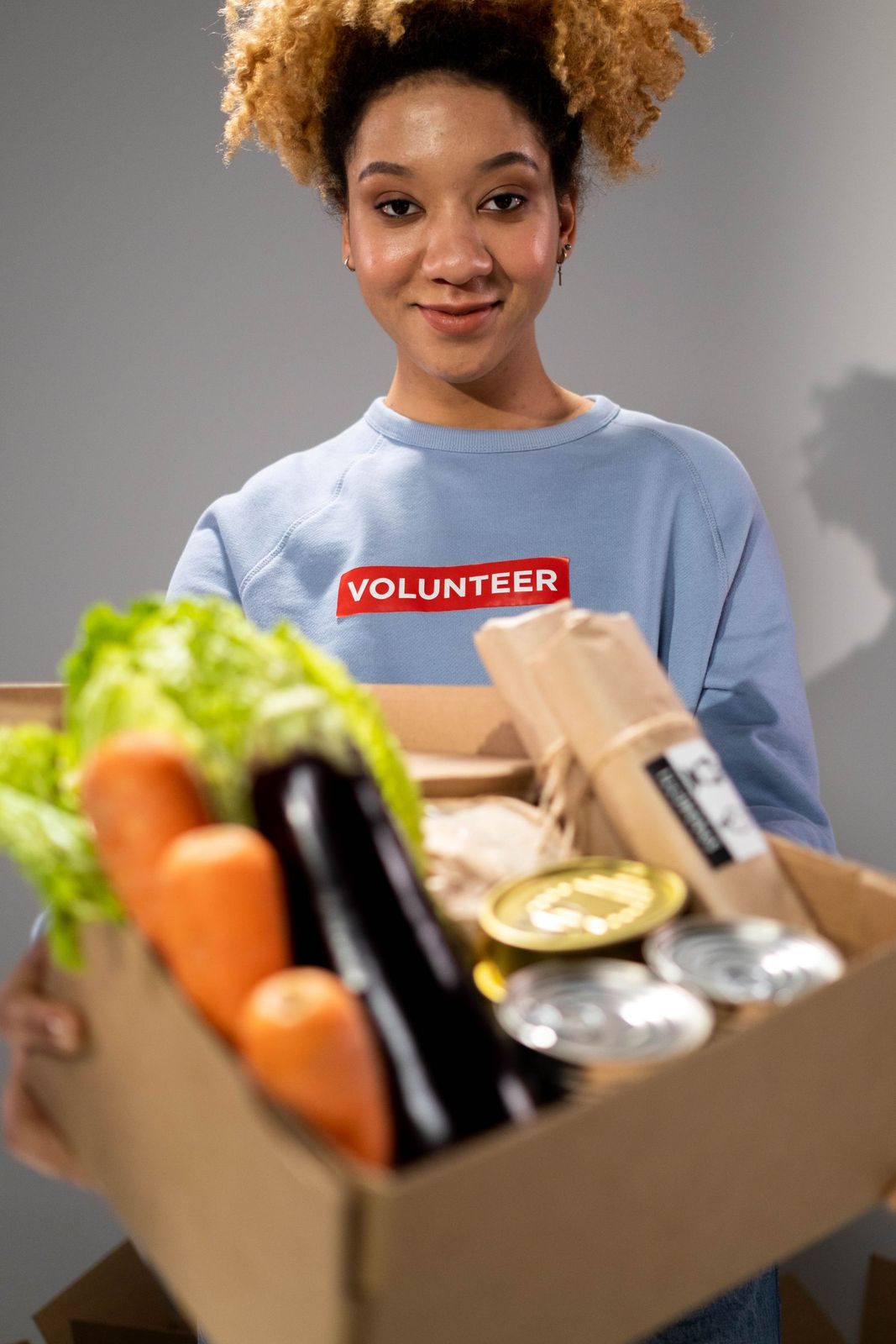 A smiling volunteer holding a box of fresh vegetables and canned goods