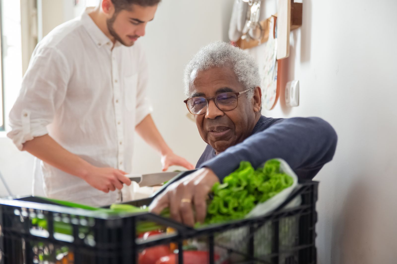 A volunteer helping an older man unpack fresh vegetables from a crate at home
